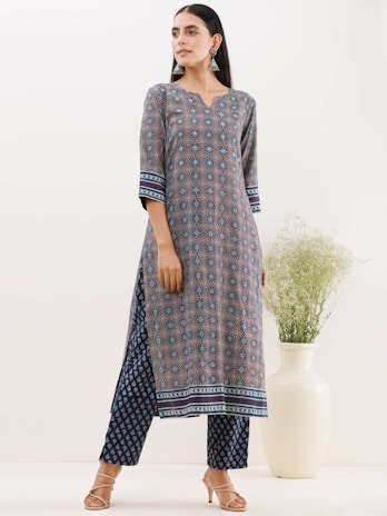 A woman is wearing a traditional patterned outfit consisting of a long kurta and matching trousers. The kurta features intricate geometric designs in shades of blue, red, and white, with strip detailing at the sleeves and hem. She stands with a slightly turned posture, adorned with large silver earrings. Her hair is styled straight and she is wearing open-toed sandals. A white vase with green foliage is placed in the background, adding a touch of nature to the setting.