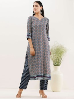 A woman is wearing a traditional patterned outfit consisting of a long kurta and matching trousers. The kurta features intricate geometric designs in shades of blue, red, and white, with strip detailing at the sleeves and hem. She stands with a slightly turned posture, adorned with large silver earrings. Her hair is styled straight and she is wearing open-toed sandals. A white vase with green foliage is placed in the background, adding a touch of nature to the setting.