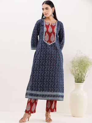 A woman wearing a traditional outfit with a navy blue patterned tunic over red patterned trousers. She has long, dark hair and is accessorized with large, ornate earrings. Behind her, there is a white vase with delicate green foliage.