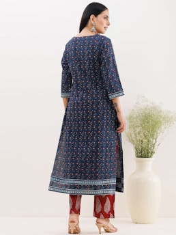 A woman stands with her back facing the camera, wearing a long blue kurta with floral patterns. The kurta is paired with red patterned trousers and beige high heels. Her long dark hair is left loose, and she wears large dangling earrings. To her side, there is a large white vase containing green foliage.