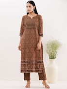 A person is wearing a long, patterned brown kurta with intricate circular and floral designs. The kurta has a distinctive border at the bottom and is paired with matching trousers. The individual is standing next to a large white vase filled with wispy green foliage. The setting is minimal with a light-colored background.