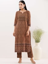 A person is wearing a long, patterned brown kurta with intricate circular and floral designs. The kurta has a distinctive border at the bottom and is paired with matching trousers. The individual is standing next to a large white vase filled with wispy green foliage. The setting is minimal with a light-colored background.