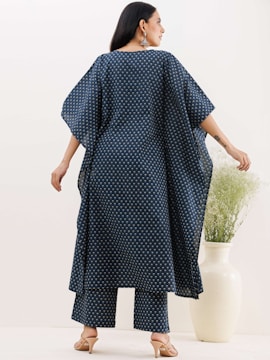 A person is wearing a dark blue, loose-fitting garment with a small, repeating pattern. The outfit consists of a long top and matching pants. The person is wearing high heels and large earrings. There is a tall white vase containing green foliage to the side.