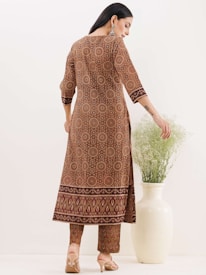 A woman is dressed in a patterned ethnic outfit, featuring circular and decorative motifs, standing with her back facing the camera. She is wearing beige high heels and her hair is styled straight. Her right hand is resting on a large white vase that contains green foliage. The background is a plain light color.