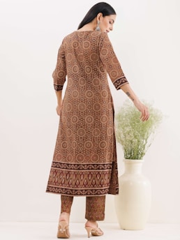 A woman is dressed in a patterned ethnic outfit, featuring circular and decorative motifs, standing with her back facing the camera. She is wearing beige high heels and her hair is styled straight. Her right hand is resting on a large white vase that contains green foliage. The background is a plain light color.