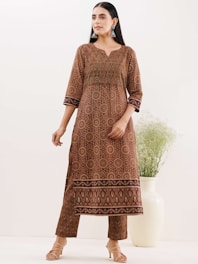 A woman wearing a brown traditional outfit with intricate patterns, including circular designs and geometric shapes. She has long, dark hair, styled straight, and is adorned with large, silver earrings. The setting is minimal, with a large white vase containing greenery in the background.