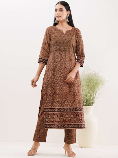A woman wearing a brown traditional outfit with intricate patterns, including circular designs and geometric shapes. She has long, dark hair, styled straight, and is adorned with large, silver earrings. The setting is minimal, with a large white vase containing greenery in the background.