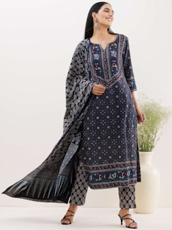 A person is wearing a traditional, dark blue ethnic outfit with intricate patterns, including floral and geometric designs. The ensemble consists of a long tunic, pants, and a matching scarf. The person is smiling and wearing large earrings, standing against a neutral background with a vase of green foliage.