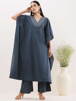 A person is wearing a blue patterned tunic and trousers ensemble. The outfit features a geometric design with a V-neckline and long flowing sleeves, paired with open-toed sandals. The person has long dark hair, large earrings, and is standing with one hand near their face. Next to them is a large white vase with green foliage.