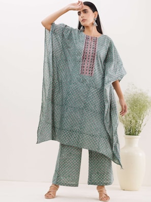 A person stands confidently wearing a flowing, patterned outfit with a geometric design. The fabric features a mix of colors, primarily muted greens and pinks. The outfit consists of a loose-fitting top and matching wide-leg pants. They are standing in a minimalist setting with a large floor vase containing green foliage in the background. The person is striking a stylish pose with one hand on their forehead, showcasing long earrings and open-toed sandals.