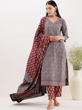 A woman wearing a traditional printed outfit with intricate patterns in tones of red, blue, and black. She stands with a draped scarf or dupatta, accessorized with earrings. She has sleek, dark hair and a calm expression. An off-white vase with green foliage is positioned on the right beside her.