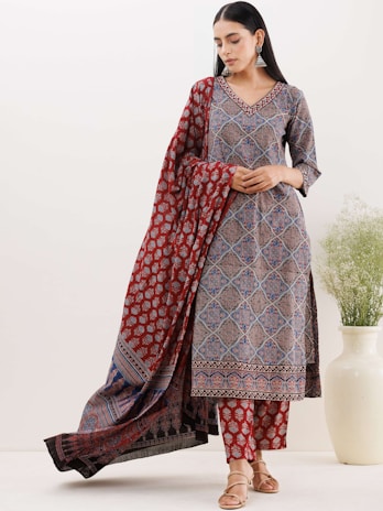 A woman wearing a traditional printed outfit with intricate patterns in tones of red, blue, and black. She stands with a draped scarf or dupatta, accessorized with earrings. She has sleek, dark hair and a calm expression. An off-white vase with green foliage is positioned on the right beside her.