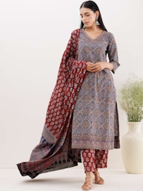 A woman wearing a traditional printed outfit with intricate patterns in tones of red, blue, and black. She stands with a draped scarf or dupatta, accessorized with earrings. She has sleek, dark hair and a calm expression. An off-white vase with green foliage is positioned on the right beside her.