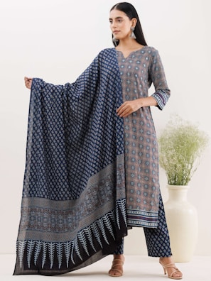 A woman is wearing a traditional outfit with intricate patterns, including a long shirt and matching pants. She is holding a matching shawl with detailed blue and black patterns, standing next to a large white ceramic vase containing green foliage. Her hair is loose and she is accessorized with large silver earrings.