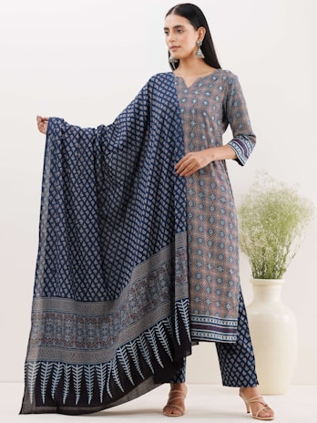 A woman is wearing a traditional outfit with intricate patterns, including a long shirt and matching pants. She is holding a matching shawl with detailed blue and black patterns, standing next to a large white ceramic vase containing green foliage. Her hair is loose and she is accessorized with large silver earrings.