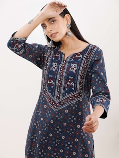 A woman is wearing a blue, patterned ethnic dress with intricate embroidery. She is posing with one arm raised above her head and the other hand near her chest. She is also wearing large, dangling earrings and appears to have a small tattoo on her wrist. The background is plain and light.