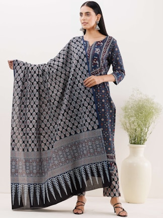 A woman stands elegantly wearing a dark blue ethnic outfit that features intricate patterns and a matching long dupatta. She holds the dupatta gracefully, showcasing its design. Her hair is neatly styled, and she wears large, traditional earrings. Next to her, there is a tall white vase with green foliage.