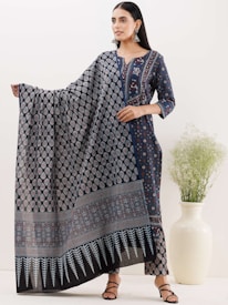 A woman stands elegantly wearing a dark blue ethnic outfit that features intricate patterns and a matching long dupatta. She holds the dupatta gracefully, showcasing its design. Her hair is neatly styled, and she wears large, traditional earrings. Next to her, there is a tall white vase with green foliage.