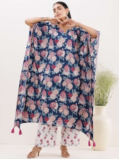 A person is modeling a loose, flowing garment with a floral pattern in shades of red, pink, and white against a dark blue background. The garment has tassels at the bottom corners. The person is posing with their arms raised and eyes closed. They are also wearing white pants with a similar floral pattern and open-toed sandals. In the background, there is a large white vase with green foliage.