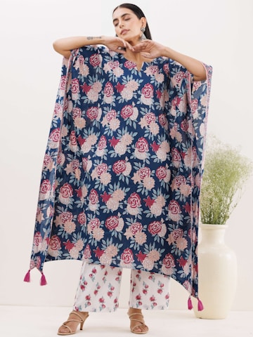 A person is modeling a loose, flowing garment with a floral pattern in shades of red, pink, and white against a dark blue background. The garment has tassels at the bottom corners. The person is posing with their arms raised and eyes closed. They are also wearing white pants with a similar floral pattern and open-toed sandals. In the background, there is a large white vase with green foliage.