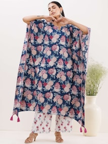 A person is modeling a loose, flowing garment with a floral pattern in shades of red, pink, and white against a dark blue background. The garment has tassels at the bottom corners. The person is posing with their arms raised and eyes closed. They are also wearing white pants with a similar floral pattern and open-toed sandals. In the background, there is a large white vase with green foliage.