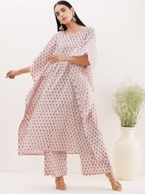 A person is wearing a flowing outfit with red floral patterns on a white background. The attire includes a loose-fitting top and matching pants. The person is standing in a neutral-colored room next to a large white vase containing green foliage. They have long dark hair and are accessorizing with large earrings and beige sandals.