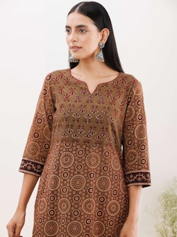 A person wearing a traditional patterned dress featuring intricate circular and floral designs in shades of brown, orange, and red. The dress has a V-neck and three-quarter sleeves. The individual is accessorized with large, ornate silver earrings and has straight, dark hair that falls past the shoulders.