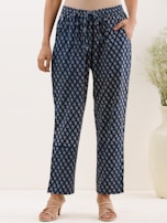 A person is wearing a pair of loose-fitting, patterned navy blue pants with a drawstring waist. The pants feature a repeating small white and red floral pattern. The person is also wearing a neutral-colored top and beige sandals with low heels. A tall white vase with greenery is visible in the background.
