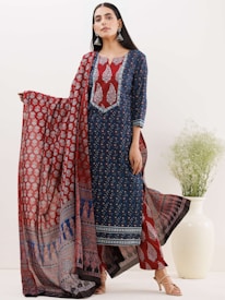 A woman is dressed in a beautifully patterned traditional outfit featuring a long blue kurta with intricate red and white designs. She holds a matching red dupatta with elaborate prints. Large, silver earrings accentuate her look, and she stands next to a white vase filled with green decorative foliage.