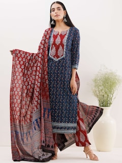 A woman is dressed in a beautifully patterned traditional outfit featuring a long blue kurta with intricate red and white designs. She holds a matching red dupatta with elaborate prints. Large, silver earrings accentuate her look, and she stands next to a white vase filled with green decorative foliage.