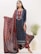A woman is dressed in a beautifully patterned traditional outfit featuring a long blue kurta with intricate red and white designs. She holds a matching red dupatta with elaborate prints. Large, silver earrings accentuate her look, and she stands next to a white vase filled with green decorative foliage.