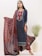 A woman is dressed in a beautifully patterned traditional outfit featuring a long blue kurta with intricate red and white designs. She holds a matching red dupatta with elaborate prints. Large, silver earrings accentuate her look, and she stands next to a white vase filled with green decorative foliage.