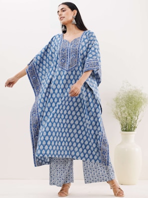 A woman is wearing a blue patterned outfit consisting of a loose-fitting tunic and matching trousers. Her hair is long and dark, and she accessorizes with large silver earrings and brown sandals. She stands next to a white ceramic vase with delicate green foliage.
