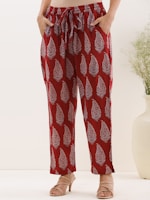 A person is standing wearing red pants with a white paisley pattern. The pants are loose-fitting with an elastic waistband and drawstring. The person is also wearing beige high-heeled sandals. There is a light, minimal background with a large white vase and a small plant.