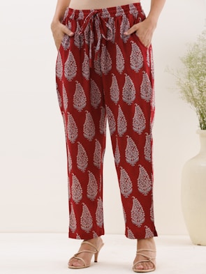 A person is standing wearing red pants with a white paisley pattern. The pants are loose-fitting with an elastic waistband and drawstring. The person is also wearing beige high-heeled sandals. There is a light, minimal background with a large white vase and a small plant.
