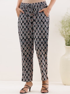 A person wearing patterned pants with a drawstring waistband. The black fabric features a repeated paisley design in light blue and maroon. The person has their hands in the pockets and is wearing black strappy sandals. A white vase with green foliage is placed on the right side of the image.