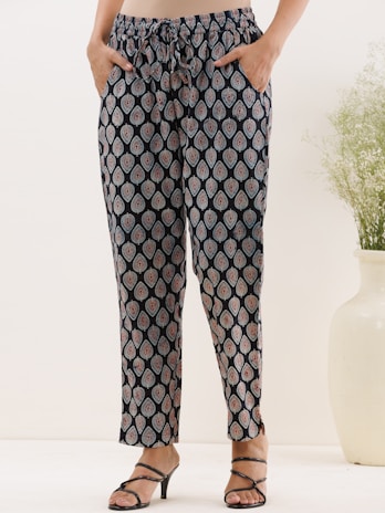 A person wearing patterned pants with a drawstring waistband. The black fabric features a repeated paisley design in light blue and maroon. The person has their hands in the pockets and is wearing black strappy sandals. A white vase with green foliage is placed on the right side of the image.