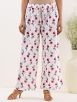 A person is wearing white loose-fitting pants with a drawstring waist. The pants are adorned with a floral pattern of red roses and green leaves. The individual is standing on a light background, wearing tan sandals with a small heel. Beside them is a large white vase containing green foliage.