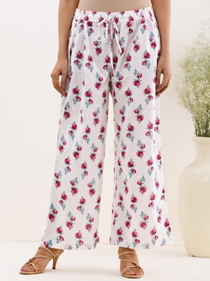 A person is wearing white loose-fitting pants with a drawstring waist. The pants are adorned with a floral pattern of red roses and green leaves. The individual is standing on a light background, wearing tan sandals with a small heel. Beside them is a large white vase containing green foliage.