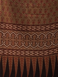 The image displays a fabric or textile with intricate patterns. The upper portion features repetitive floral motifs in red on a beige background. Below it, there is a segment with zigzag patterns in shades of brown. This is followed by a section with symmetrical, ornamental designs in beige and dark brown. The bottom-most part showcases vertical elements, resembling leaves or feathers, in red and black.