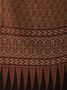 The image displays a fabric or textile with intricate patterns. The upper portion features repetitive floral motifs in red on a beige background. Below it, there is a segment with zigzag patterns in shades of brown. This is followed by a section with symmetrical, ornamental designs in beige and dark brown. The bottom-most part showcases vertical elements, resembling leaves or feathers, in red and black.