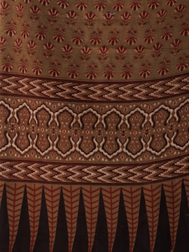 The image displays a fabric or textile with intricate patterns. The upper portion features repetitive floral motifs in red on a beige background. Below it, there is a segment with zigzag patterns in shades of brown. This is followed by a section with symmetrical, ornamental designs in beige and dark brown. The bottom-most part showcases vertical elements, resembling leaves or feathers, in red and black.