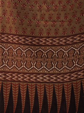 The image displays a fabric or textile with intricate patterns. The upper portion features repetitive floral motifs in red on a beige background. Below it, there is a segment with zigzag patterns in shades of brown. This is followed by a section with symmetrical, ornamental designs in beige and dark brown. The bottom-most part showcases vertical elements, resembling leaves or feathers, in red and black.
