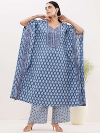 A person wearing a flowing blue traditional outfit with a detailed pattern. The attire includes a long tunic and wide-legged pants with matching designs. The background is minimal, featuring a neutral wall and a plant in a white vase. The person wears earrings and sandals.