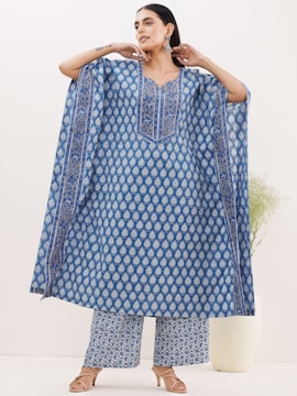A person wearing a flowing blue traditional outfit with a detailed pattern. The attire includes a long tunic and wide-legged pants with matching designs. The background is minimal, featuring a neutral wall and a plant in a white vase. The person wears earrings and sandals.