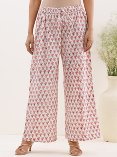A person standing wearing loose, white wide-legged pants with a red floral pattern. The person is also wearing beige sandals. In the background, there's a large white vase containing green foliage, and the setting appears to be indoors with a simple and minimalistic decor.