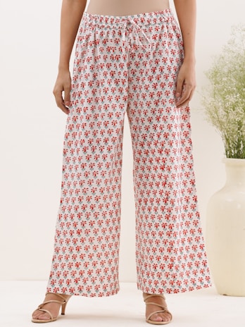 A person standing wearing loose, white wide-legged pants with a red floral pattern. The person is also wearing beige sandals. In the background, there's a large white vase containing green foliage, and the setting appears to be indoors with a simple and minimalistic decor.