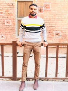A stylish man wearing a cozy knit sweater and jeans, standing against a rustic brick wall.