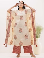 A person is wearing a traditional outfit that features a cream-colored kurta with floral patterns and intricate embroidered designs on the border. The ensemble includes matching pants and beige heeled sandals. The person is accessorizing with large earrings and has long, straight hair. In the background, there is a large white vase with green foliage.
