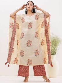 A person is wearing a traditional outfit that features a cream-colored kurta with floral patterns and intricate embroidered designs on the border. The ensemble includes matching pants and beige heeled sandals. The person is accessorizing with large earrings and has long, straight hair. In the background, there is a large white vase with green foliage.